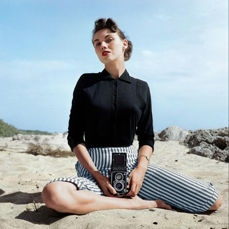 A woman sits on a sandy beach holding a vintage camera. She wears a black long-sleeve shirt and striped pants. Her hair is styled in an elegant, retro fashion. The sky is clear with scattered clouds, and rocks are visible in the background.