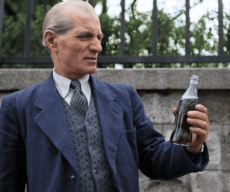 An older man in a suit and tie holds a Coca-Cola bottle, examining it closely. He stands in front of a stone wall with a black iron fence and trees visible in the background.