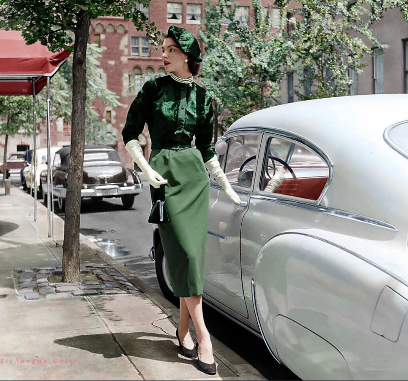 A woman in a vintage green dress and hat stands next to a classic car on a city street. She wears long gloves and pearl accessories. The background shows buildings, trees, and parked cars, creating a mid-20th century urban atmosphere.