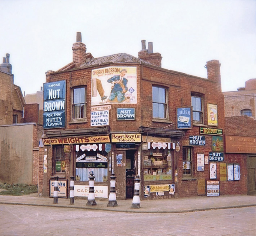 A brick corner shop with numerous vintage advertisements for tobacco and cigarettes, including "Nut Brown" and "Cherry Blossom." The shop displays various signs and posters on the walls and windows.