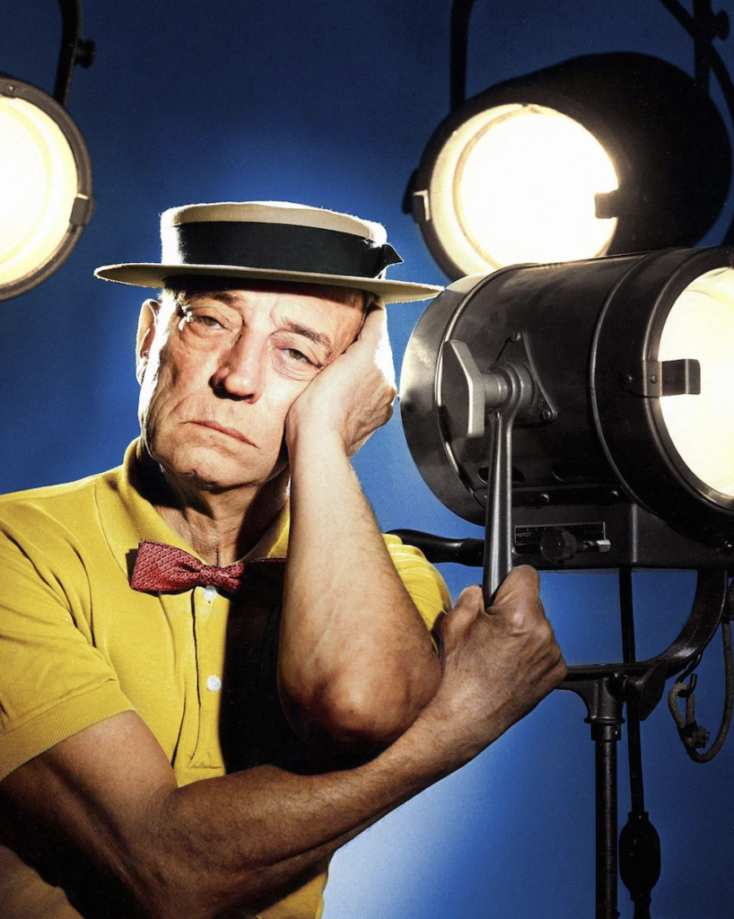 An elderly man in a yellow shirt, red bow tie, and straw boater hat rests his head on his hand with a thoughtful expression. He leans against a large studio spotlight, with two more shining in the background.