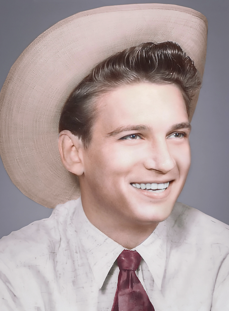 A man with a wide smile is wearing a light cowboy hat, a light shirt, and a red tie. He has short, neatly styled hair and is looking slightly to the side. The background is a plain gray.