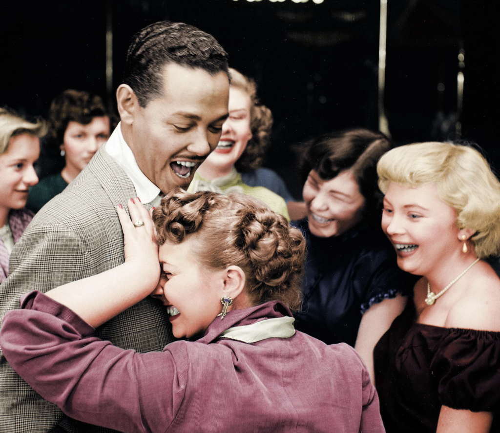 A group of people laughing joyfully at a social gathering. A man in a suit and tie smiles broadly, while a woman in a purple outfit leans on his shoulder, laughing. Others around them are also sharing in the amusement.