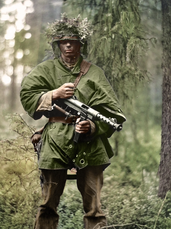 A soldier stands in a forest, wearing a green poncho and a helmet adorned with foliage. He holds a gun, with trees and undergrowth surrounding him, blending into the woodland environment.