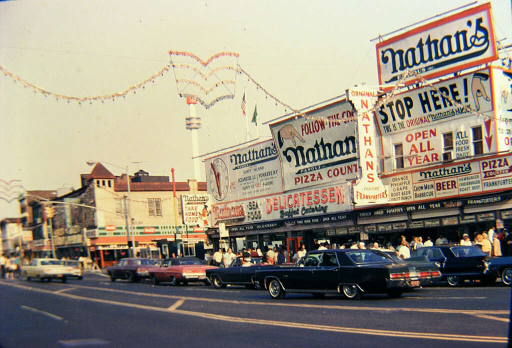 Vintage street scene featuring Nathan's Famous hot dog stand, with colorful signage and a crowd of people. Classic cars are parked along the road, and festive decorations are strung overhead, creating a lively, nostalgic atmosphere.