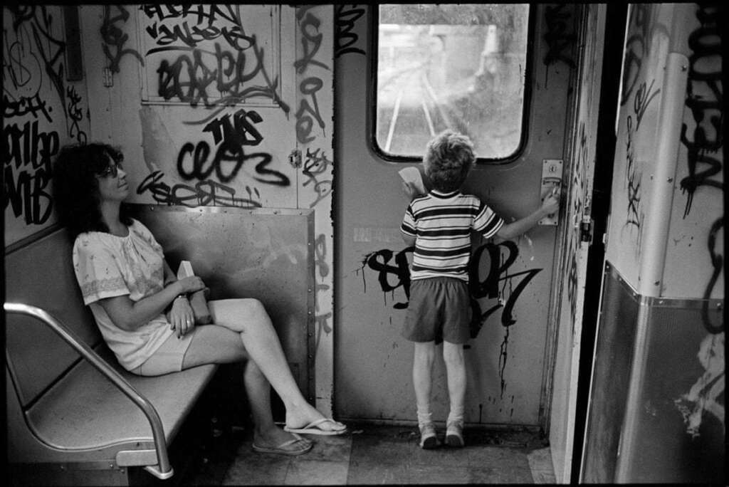 A woman sits smiling on a subway seat, holding a book. A child stands near the door, facing and looking out the window. The subway car walls are covered in graffiti.