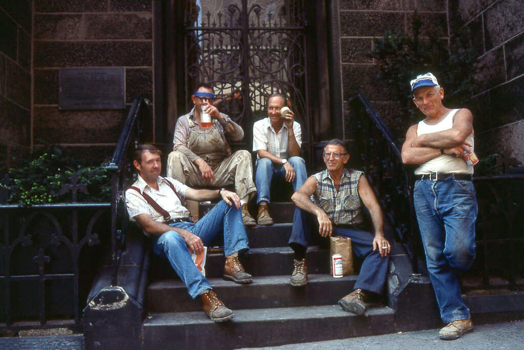 Five men in casual work attire sit and stand on stone steps in front of an ornate metal gate. Four hold drinks, and one stands with crossed arms. Sunlight casts shadows, suggesting a relaxed break.