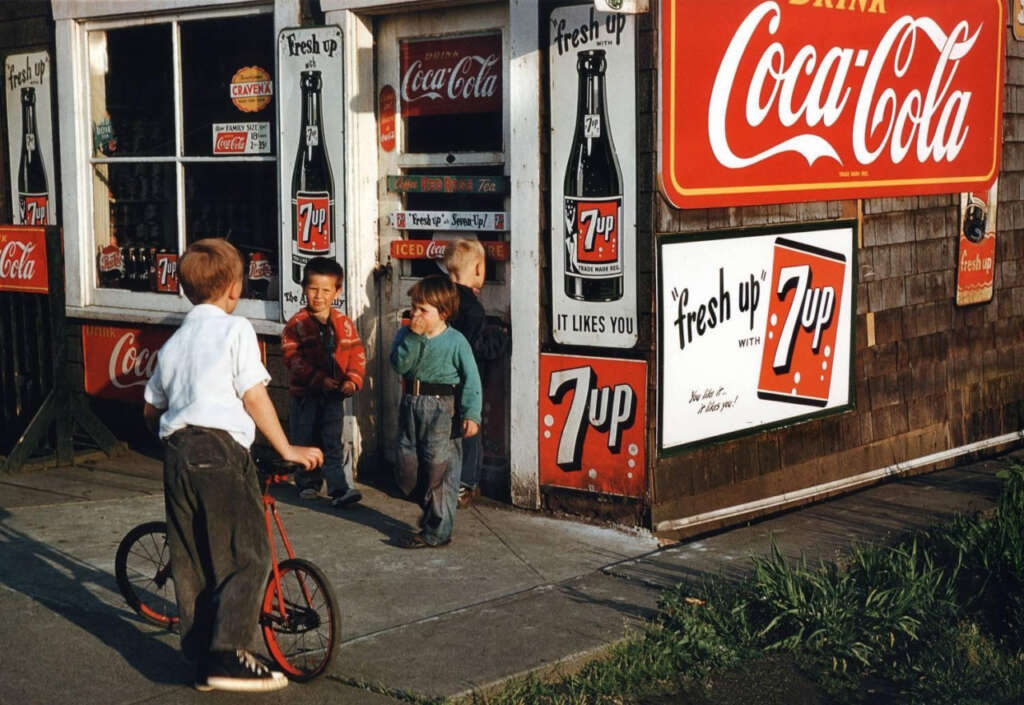 Children stand near a corner store with prominent Coca-Cola and 7-Up signs. One child holds a tricycle, while others chat and laugh. The store's wooden exterior is adorned with vintage soda advertisements. The scene captures a nostalgic, lively moment.