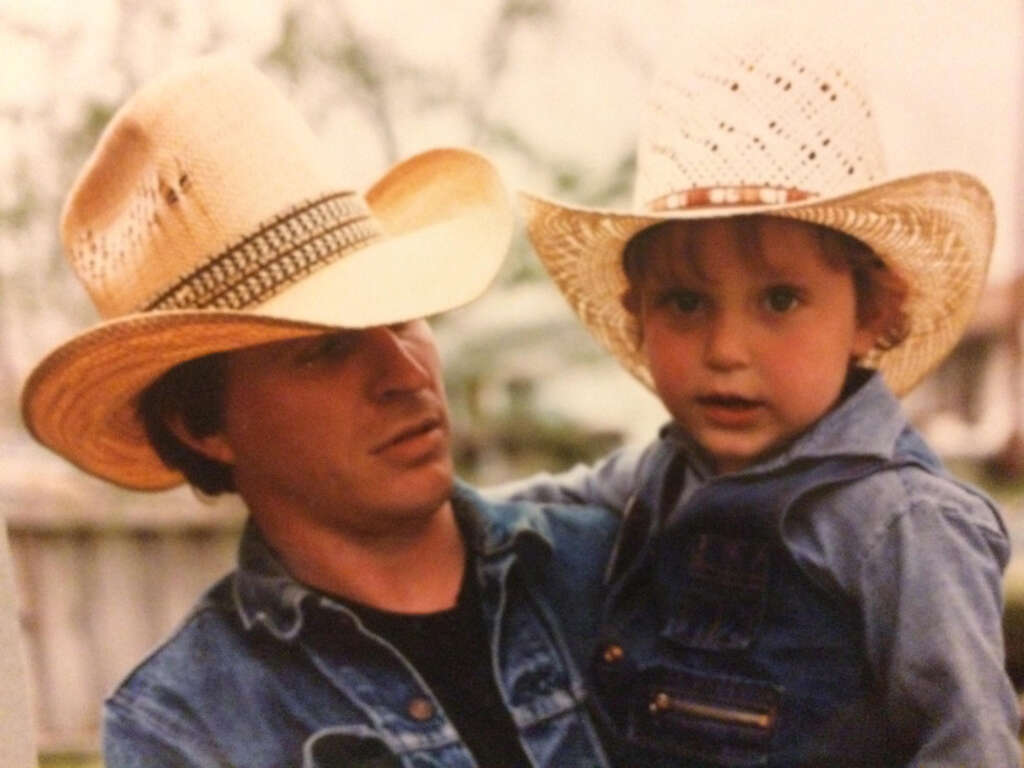 A man wearing a straw cowboy hat and denim jacket holds a young child dressed in a similar straw hat and denim outfit. They are outdoors, with blurred greenery in the background, both looking towards the camera.