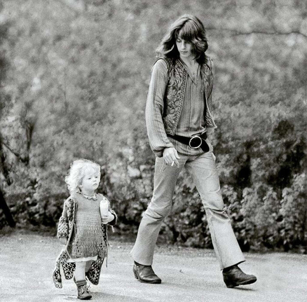 A black and white photo of an adult and a child walking outdoors. The adult has long hair and wears a patterned vest, striped shirt, and bell-bottom pants. The child, with curly hair, wears a knitted dress and cardigan. Trees form the background.