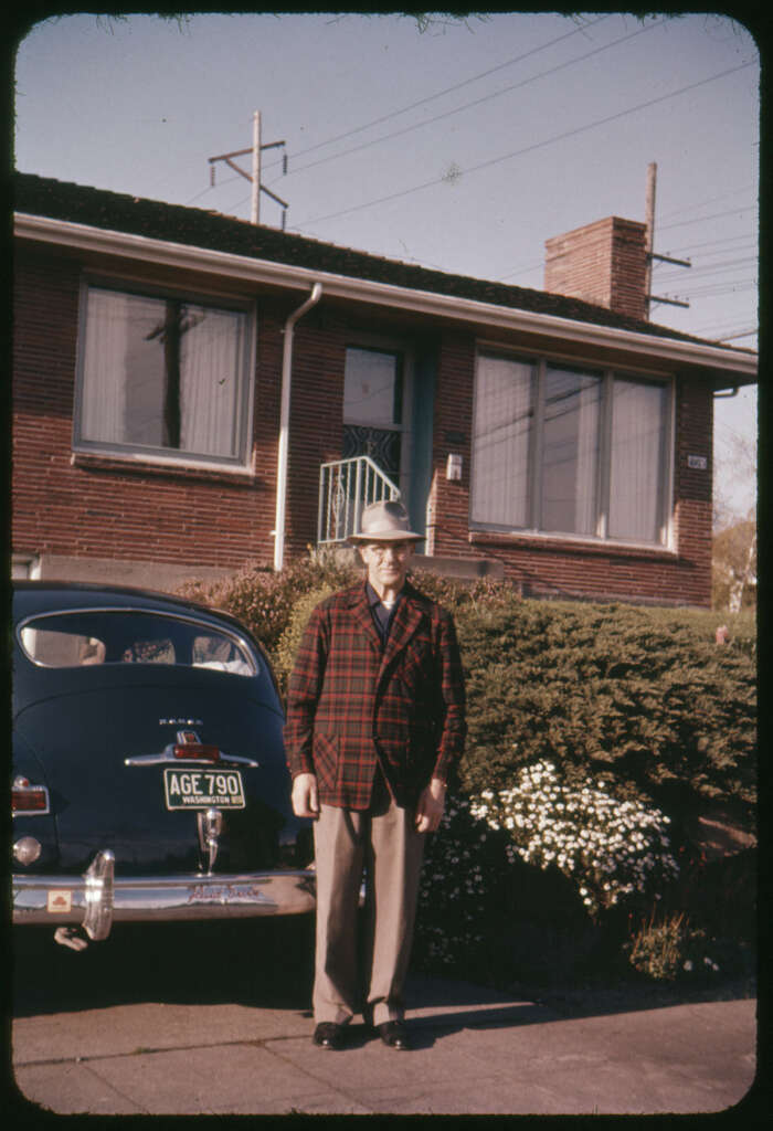 A man in a plaid jacket and hat stands beside a vintage car with a visible license plate. He's in front of a brick house with large windows and surrounded by bushes and white flowers. Power lines are in the background.