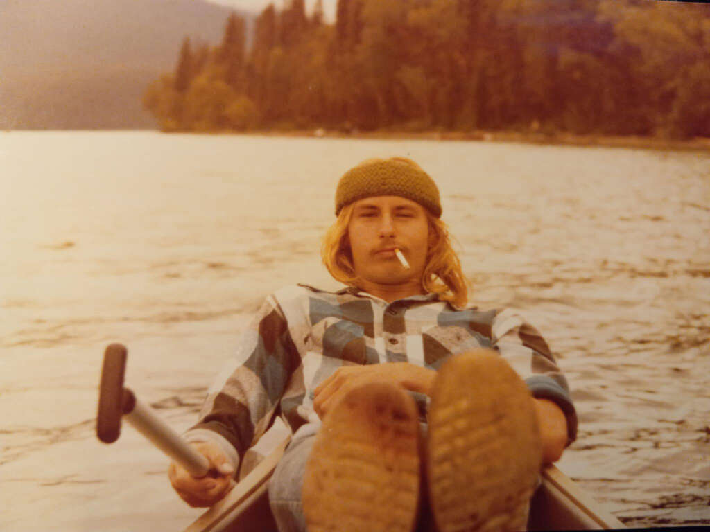 Person reclines in a rowboat on a lake, holding an oar, with a cigarette in their mouth. They wear a plaid shirt, jeans, and a knit cap. Trees and hills line the shore in the background under an overcast sky.