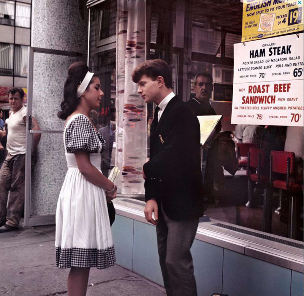 A young woman in a checkered dress and headband stands talking to a young man wearing a suit jacket. They are outside a diner with food signs in the window, and people are passing by on the street.