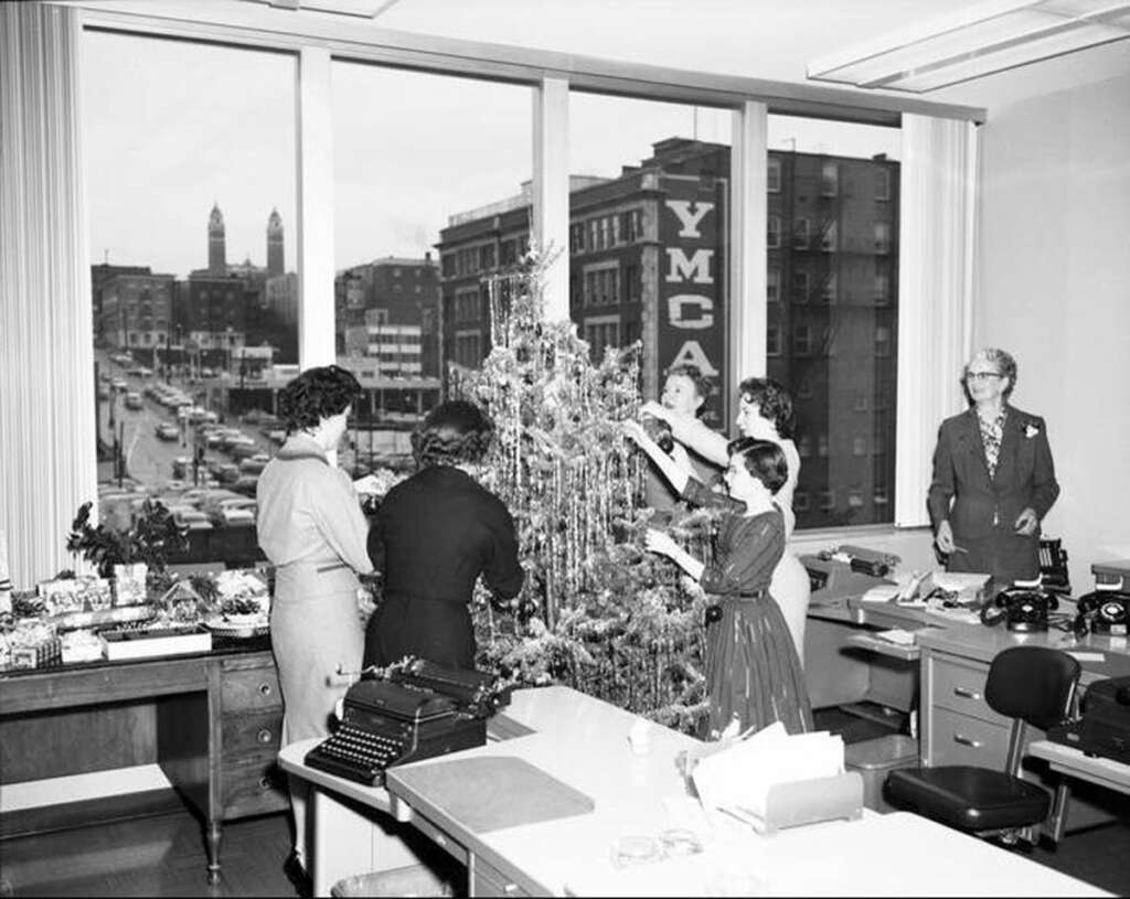 A group of women decorate a Christmas tree in an office with large windows. In the background, the cityscape features a prominent "YMCA" sign. The room has desks, typewriters, and office chairs.