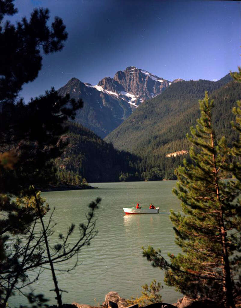 A serene mountain lake surrounded by pine trees, with a small boat carrying two people on the calm water. In the background, majestic snow-capped mountains rise under a clear blue sky.