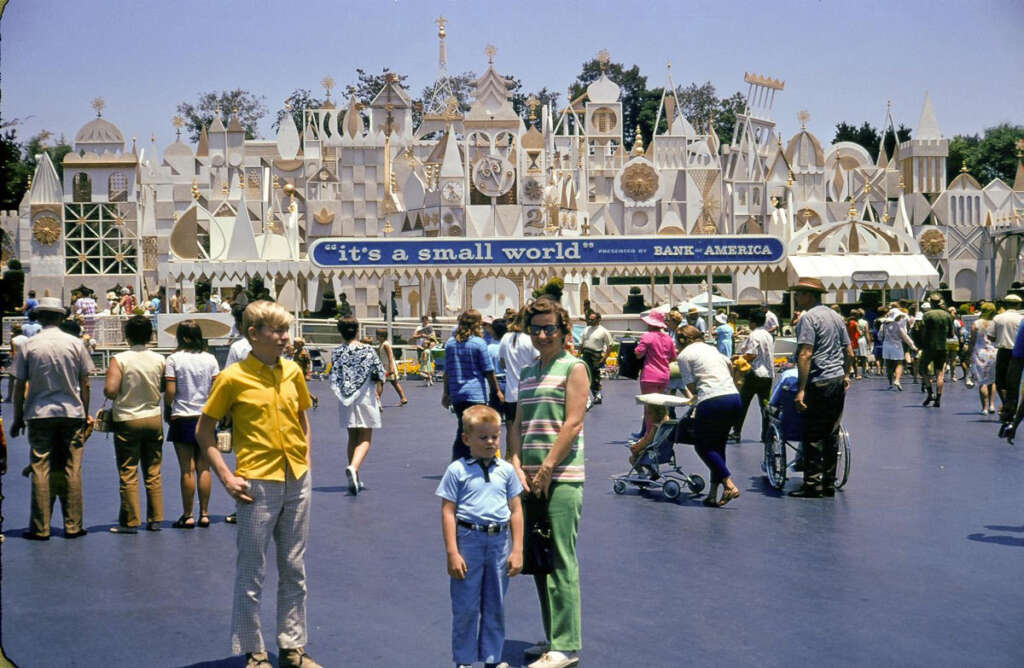 A group of people stands in front of the "it's a small world" attraction at a theme park. The building features whimsical, abstract architecture. A woman and two children pose in the foreground while other visitors walk around. It's a sunny day.