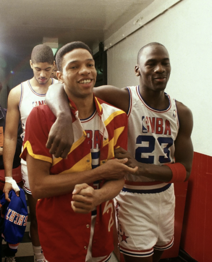 Three basketball players walk down a hallway. The player in the center wears a red and yellow jacket and smiles, while the player on the right, in a white NBA jersey with "23" and a red wristband, places an arm around him.