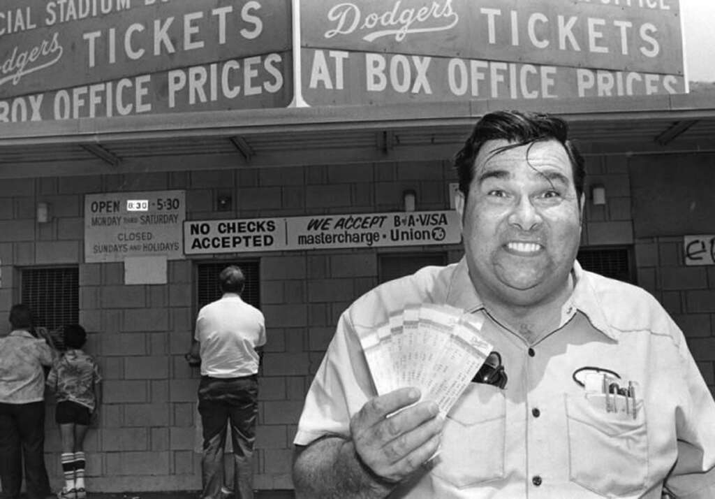 A man smiling and holding several tickets stands in front of a ticket booth. The sign above reads "Dodgers Tickets" with prices and policies. A few people are lined up at the booth. The setting appears to be at a stadium entrance.