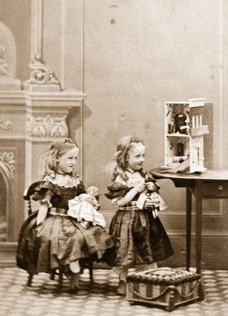 Two young girls in Victorian-era dresses sit on chairs, playing with a dollhouse on a table. Each holds a doll. The room features ornate decor and a patterned carpet.