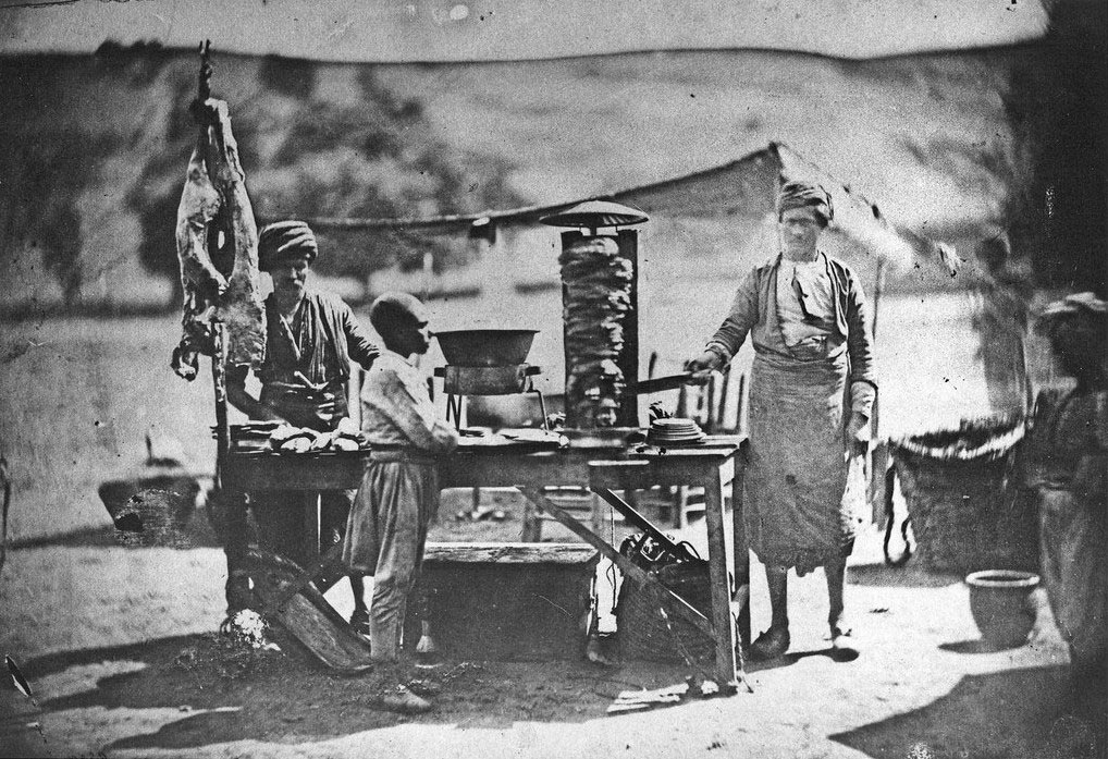 A vintage photo shows two men and a child at an outdoor food stand. Meat hangs on one side, and a vertical rotisserie is at the center. The background features a rural setting with hills and a small tent.