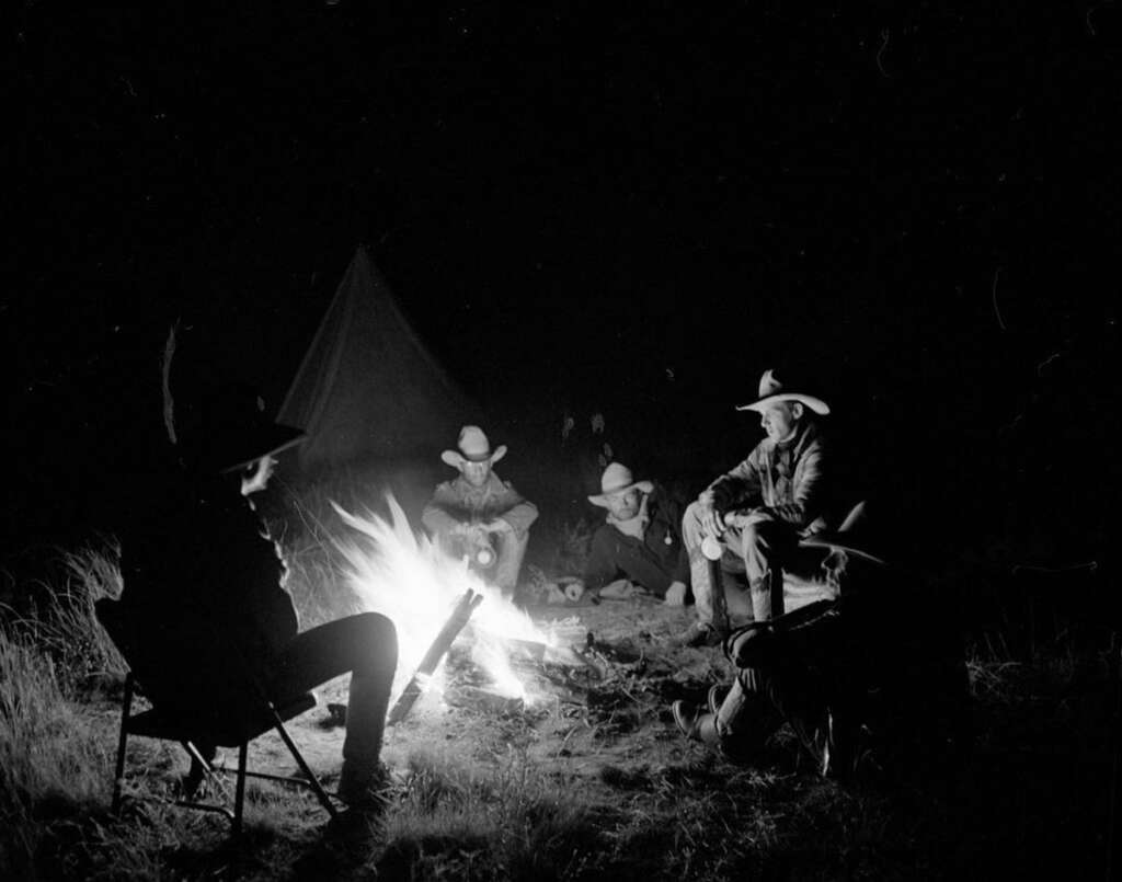 A group of cowboys with hats gathers around a campfire at night. They sit on the ground and in chairs, illuminated by the fire's glow. A tent is visible in the background amid the darkness.