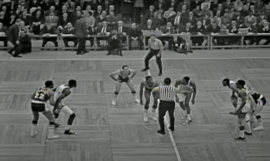 A vintage black-and-white photo of a basketball game tip-off. Players from opposing teams stand ready around the center circle as the referee holds the ball. A crowd watches from the background, seated closely along the court's sidelines.