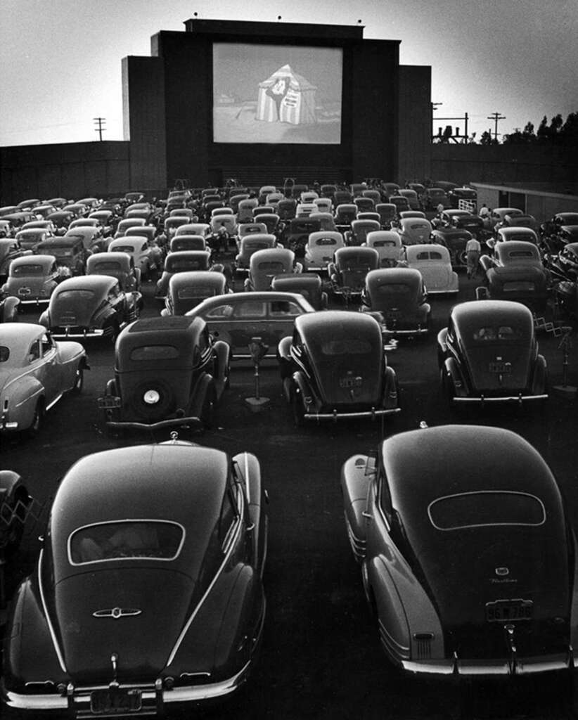 A black and white photo of a crowded drive-in theater filled with classic cars facing a large screen. The screen displays a movie scene of a person in a white cloak. Trees and power lines are visible in the background.