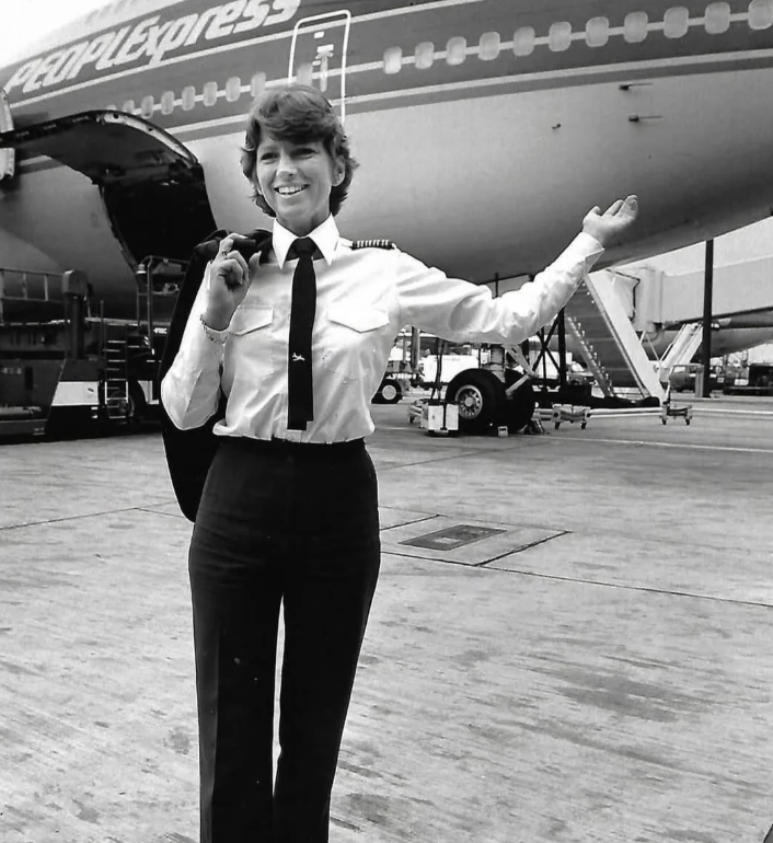 A pilot in uniform stands confidently on the tarmac, smiling with one arm raised toward a People Express airplane behind her. She holds her jacket over her shoulder, and the airplane door is open with steps leading up to it. The image is in black and white.