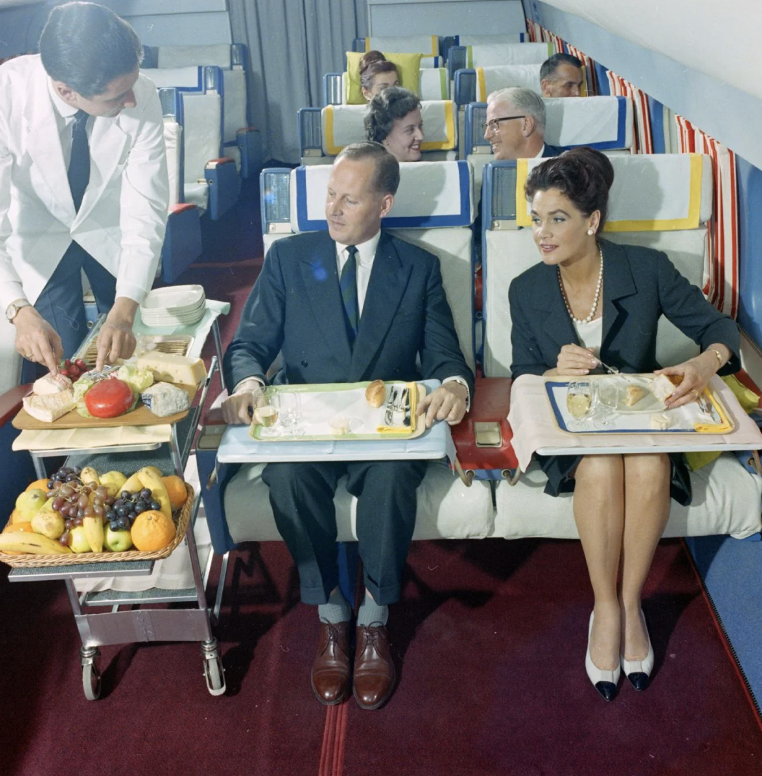 A flight attendant serves a couple seated in a vintage airplane's first-class cabin. They have trays with food on their laps. Other passengers are seated behind them. A trolley with assorted fruit and bread is beside the attendant.