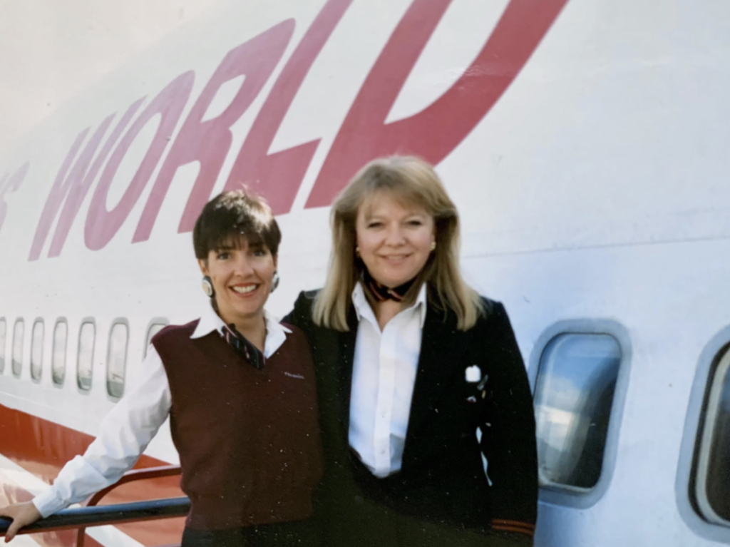 Two flight attendants in uniform stand smiling in front of an airplane with red lettering partially visible, spelling "WORLD." The aircraft appears to be a commercial jetliner, and the sky is clear.