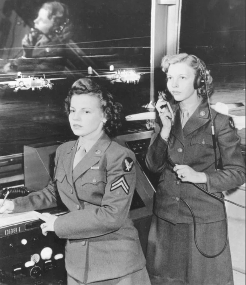 Two women in military uniforms work at a radar station during World War II. One writes on a notepad, while the other listens intently with a headset, standing near equipment. A large radar screen with aircraft models is visible behind them.