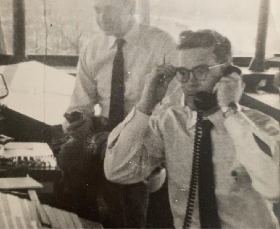 Two men in a retro office setting. One is sitting at a desk with a phone to his ear and adjusting glasses. Papers and office supplies are scattered around. The standing man looks on, holding something in his hand. Both wear white shirts and ties.