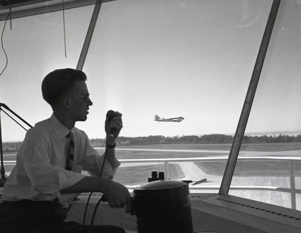 A black and white photo shows a person in a tower holding a microphone, observing a plane taking off or landing on a runway. The windows reveal an airfield with trees in the background. The person is wearing glasses and a tie.