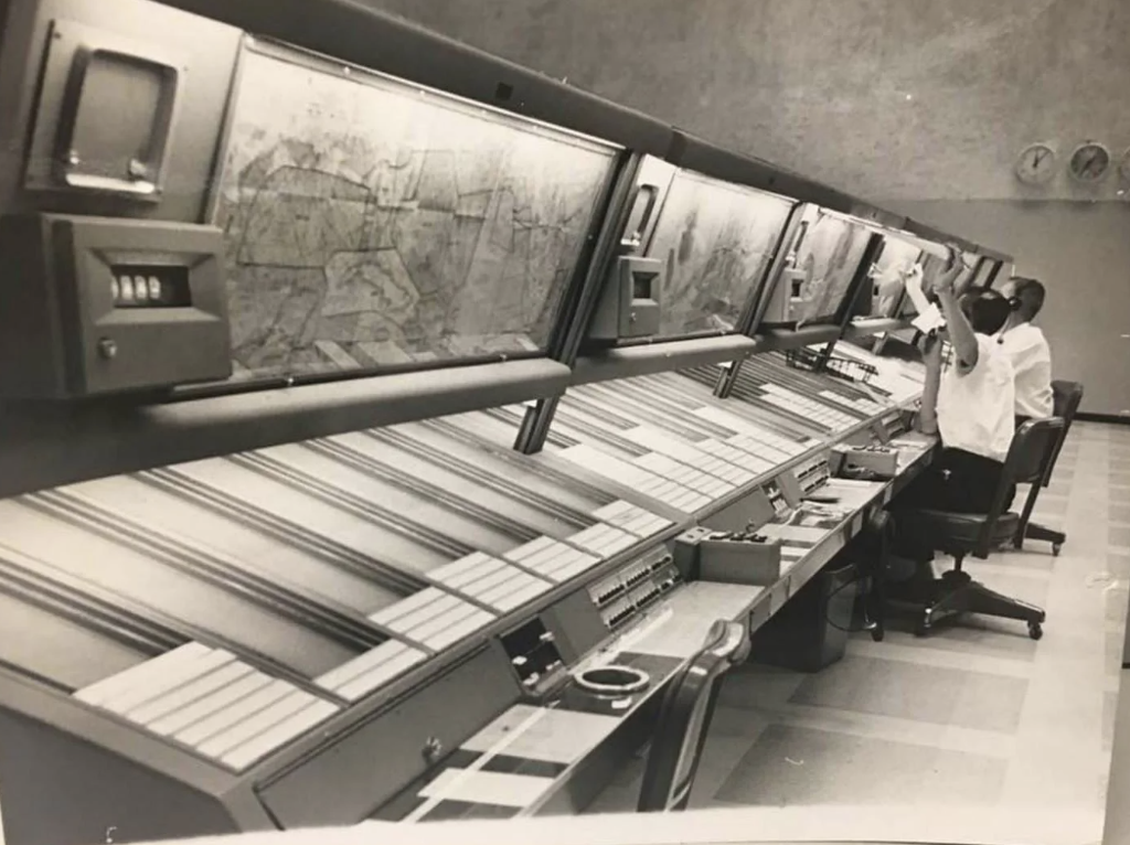 A vintage black and white photo shows a man in a suit operating a large, angled control panel with buttons and dials. The panel has illuminated screens above with maps or charts. The setting is a retro control room.