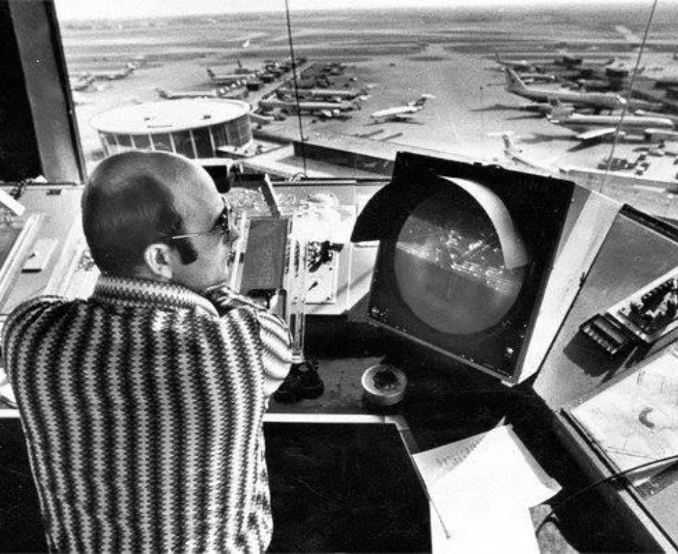 An air traffic controller wearing a patterned sweater is seated in a control tower, viewing a radar screen. The airport tarmac is visible through the window, showing several airplanes parked and taxiing.