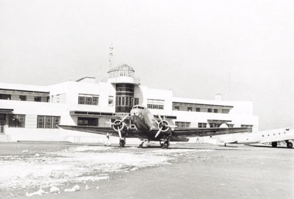 A vintage black and white photograph of a plane parked on the tarmac in front of a mid-20th century airport terminal. The building has large windows and a central tower. Scattered snow patches are visible on the ground.