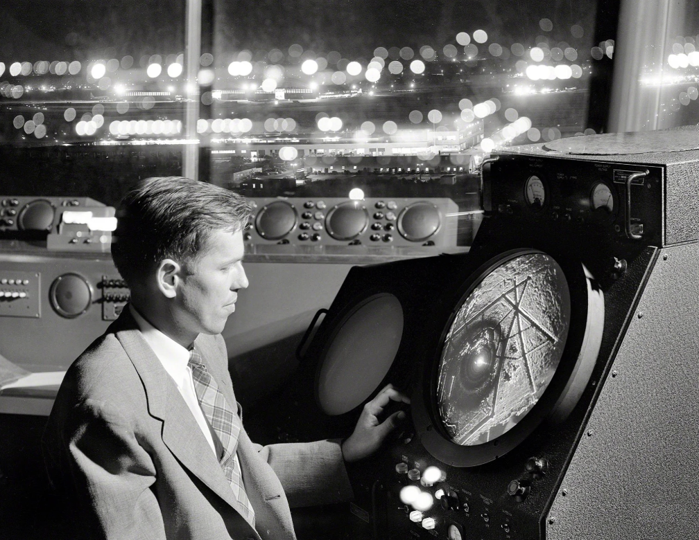 A person in a suit sits at a radar console in a control room. The large round screen displays data. Blurred lights from a cityscape are visible through the window in the background.