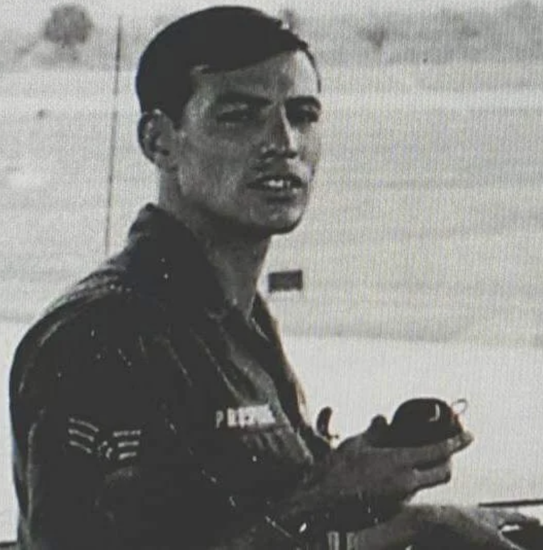 A young man in military attire is holding an object in his hand. He stands outdoors with a barren landscape in the background. The image is black and white, focusing on his serious expression and uniform details.