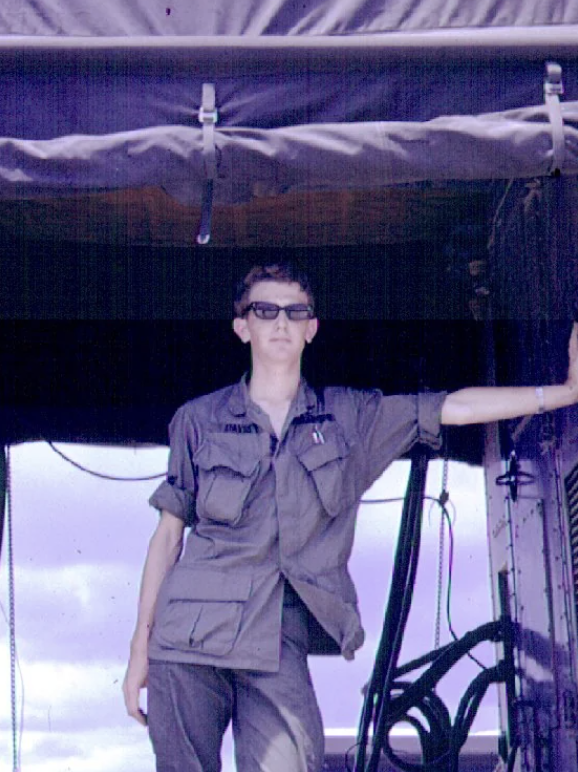 A person wearing a military-style uniform stands outdoors, leaning against a dark structure. They are wearing sunglasses and have short hair. The background shows a partly cloudy sky.