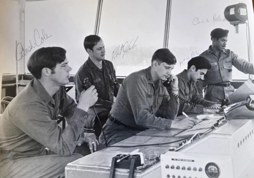 Five men in military uniforms are seated at a control console. One holds a microphone, another is on the phone, and a third is writing. Two others are observing. Various controls and communications equipment are visible on the console.