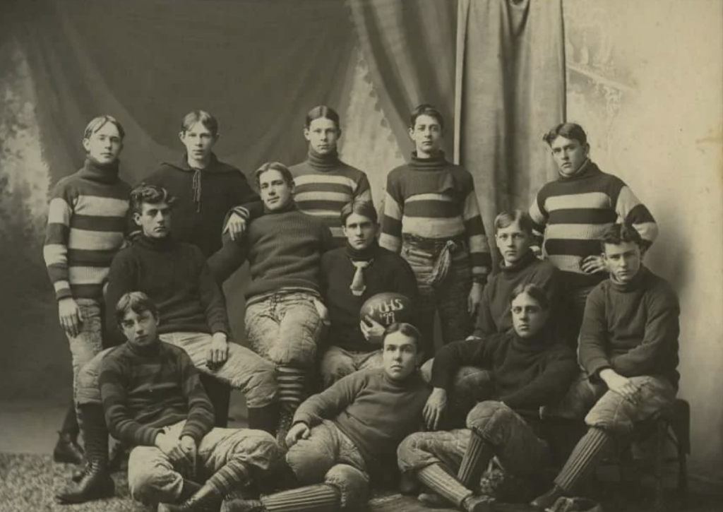 A vintage black and white photograph of a football team with thirteen young men. They're wearing striped sweaters and padded pants, posed together against a draped backdrop. Their expressions are serious, capturing a sense of camaraderie.