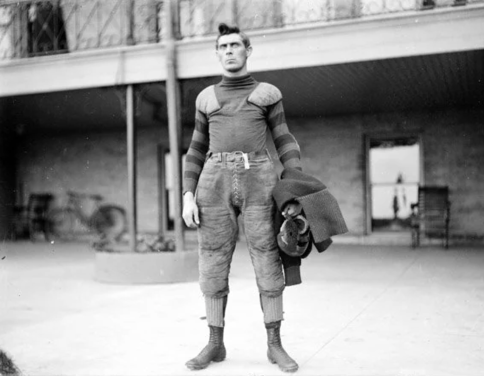 A vintage photo of a man in an early 20th-century American football uniform. He is holding a helmet and gear, standing on a porch with a bicycle in the background. The outfit includes a thick sweater and padded pants.