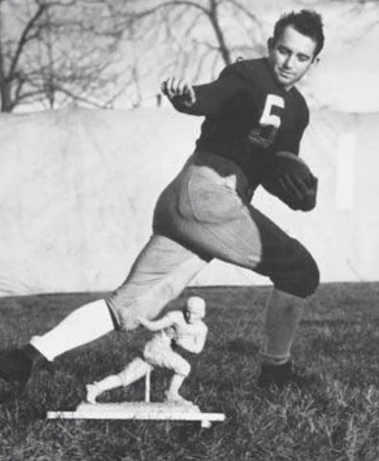 Black and white photo of a football player in a dark jersey with the number 5, posing with a football. He stands next to a trophy on the ground, mimicking its stance. Trees and a blank backdrop are in the background.