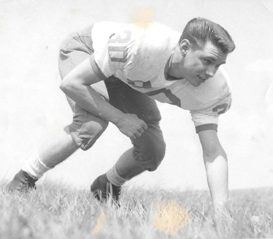 Black and white photo of a football player in a three-point stance on grass. He wears a white jersey with the number 30 and dark pants. The sky is visible in the background.