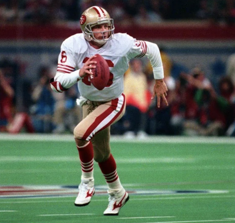 A football player in a red and white uniform and helmet runs with the ball during a game. The uniform has the number "16" on it. The action takes place on a field with blurred spectators in the background.