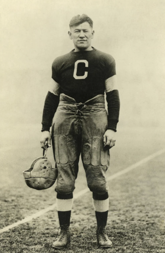 A sepia-toned vintage photo of a man in early 20th-century football gear. He stands on a field, wearing a dark jersey with a large "C" on the chest, padded pants, and holds a leather helmet in his right hand.