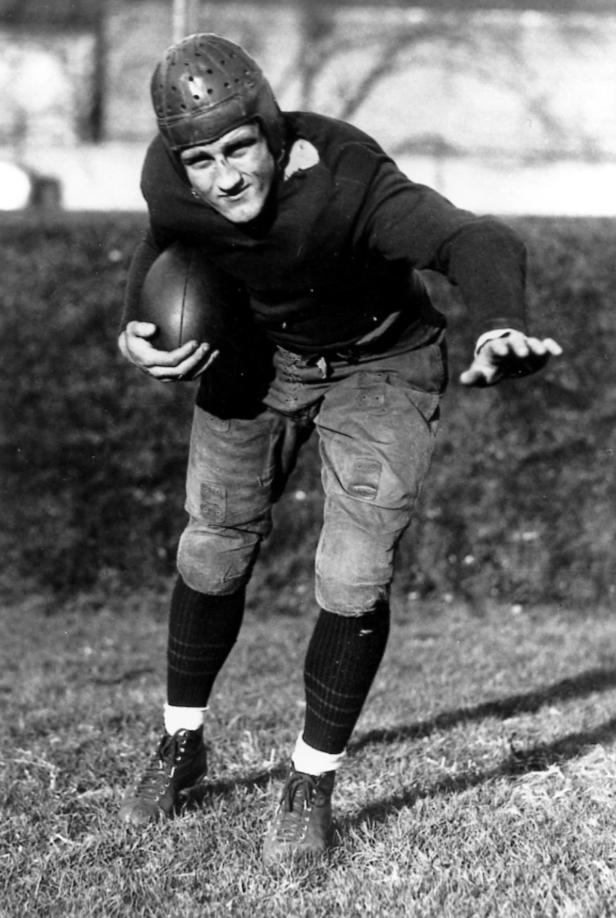 A vintage black and white photo of an American football player in early 20th-century gear. He's wearing a leather helmet and padded uniform, holding a football tucked under one arm, with the other arm outstretched in motion on a grassy field.