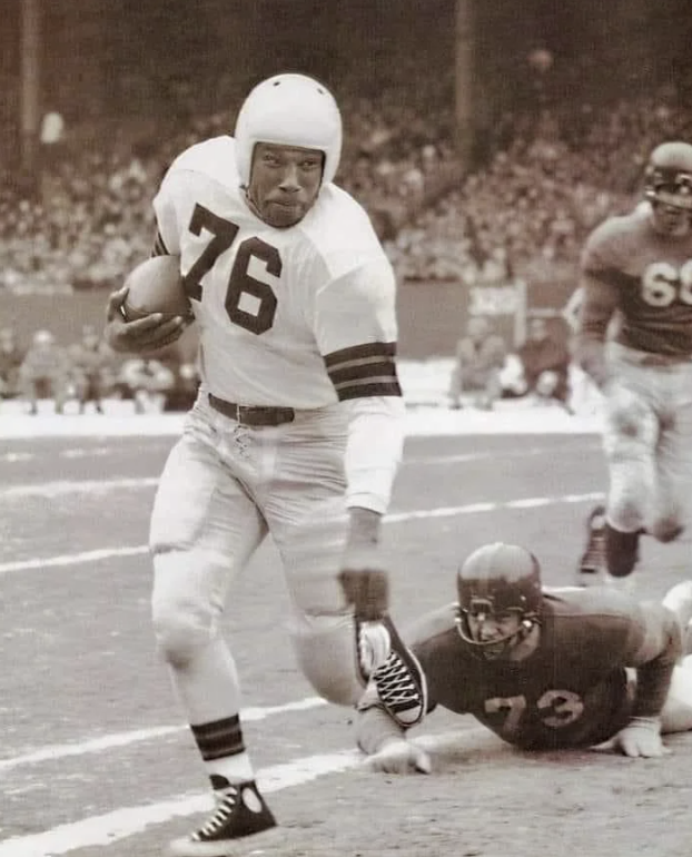 A vintage football scene showing a player in a white uniform with the number 76 running with the ball. Another player in a dark uniform is on the ground reaching for him. A crowd watches in the stadium background.