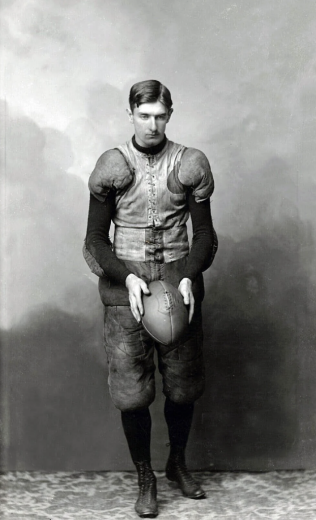 A vintage black-and-white photo of a man in early 20th-century football gear, holding a football. He wears padded attire and lace-up boots, standing in front of a plain, textured backdrop.