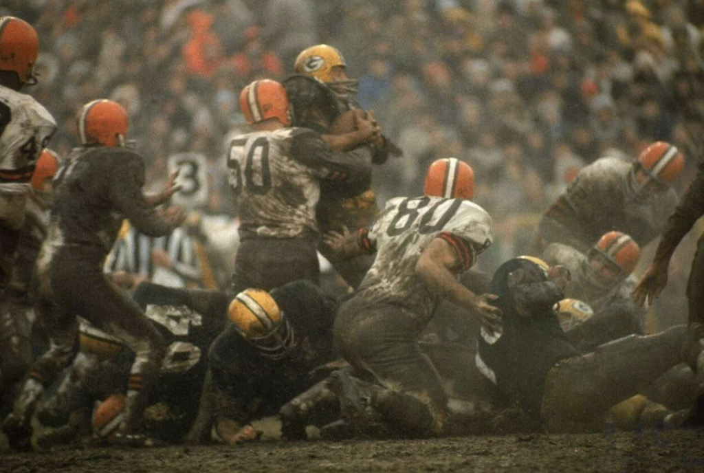 A muddy football game with players in orange and white uniforms tackling a player in a green and yellow uniform. The field is crowded and the scene is action-packed under overcast skies.